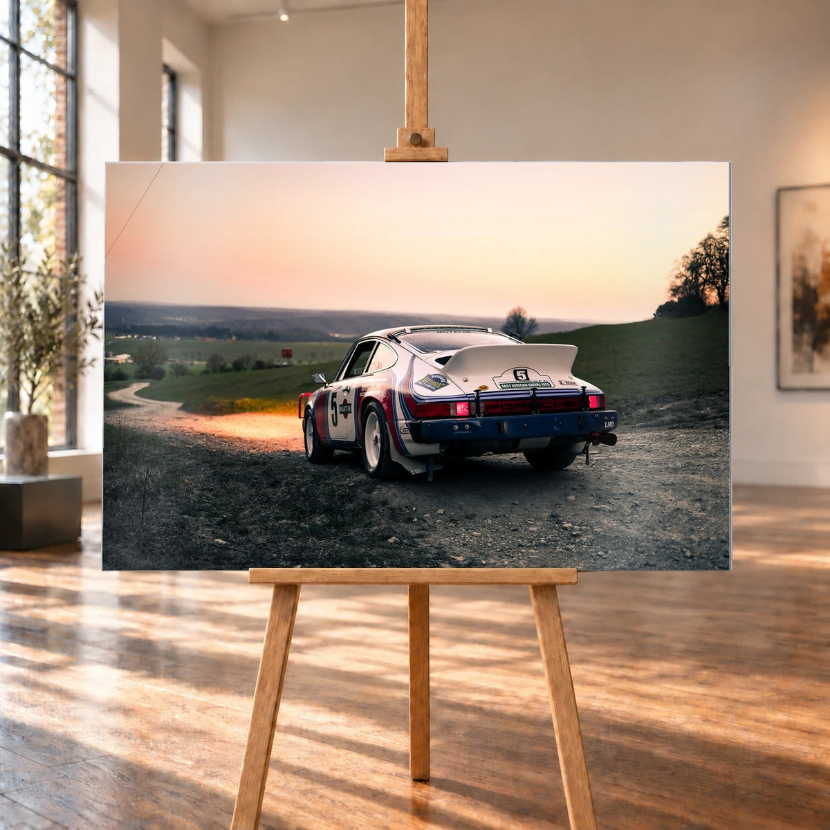 Der Porsche 911 Safari steht von hinten auf einer Anhöhe im Abendrot; weiter Himmel und orangefarbene Horizontlinie öffnen das Bild. Motiv „Fast Motion“ als Alu gebürstet-Wandbild, auf einer Holzstaffelei in einem hellen Galerieraum.
