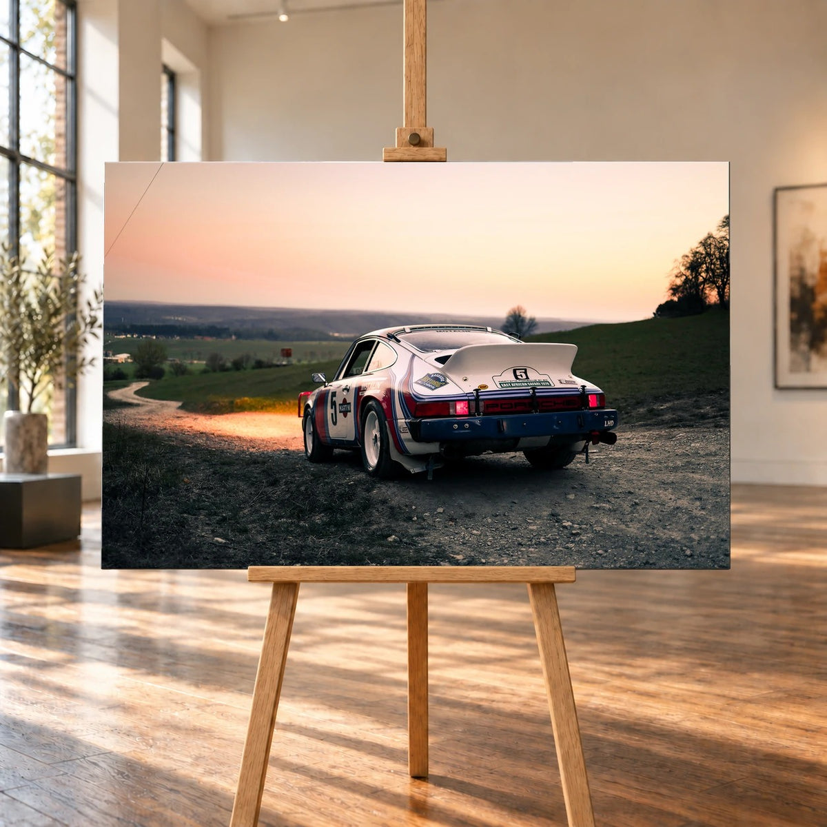 Der Porsche 911 Safari steht von hinten auf einer Anhöhe im Abendrot; weiter Himmel und orangefarbene Horizontlinie öffnen das Bild. Motiv „Fast Motion“ als Leinwand-Wandbild, auf einer Holzstaffelei in einem hellen Galerieraum.