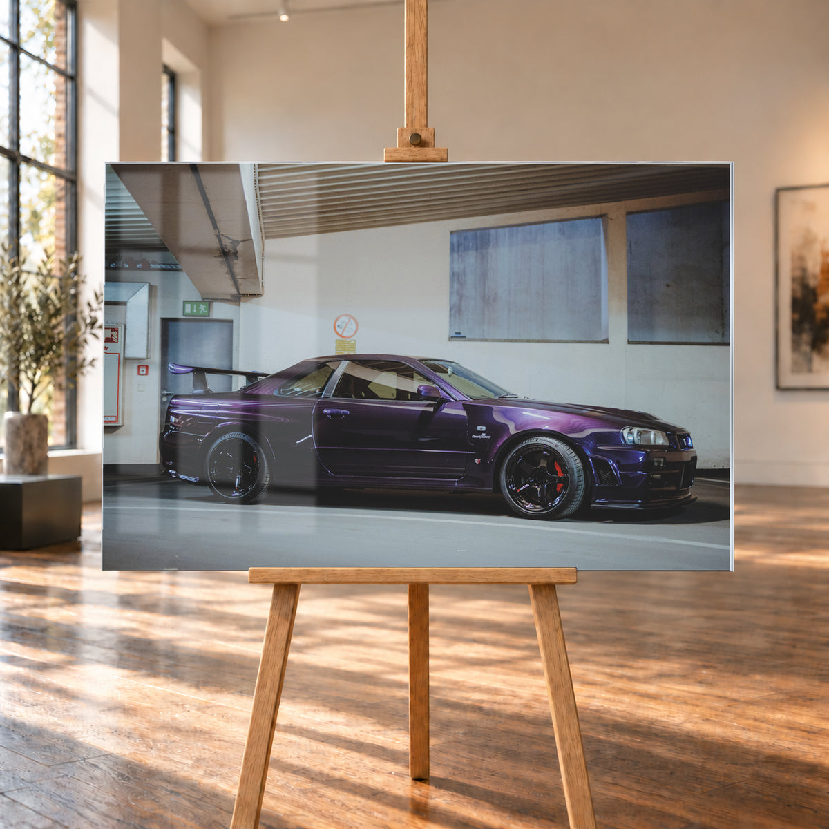 Violetter Nissan Skyline R34, nahezu seitlich mit der Front nach rechts, in einem hellen Parkgaragen-Innenraum; sichtbar sind schwarze Räder, rote Bremssättel und ein großer Heckflügel; Visualisierung als Acrylglas-Wandbild, auf einer Holzstaffelei in einem hellen Galerieraum.