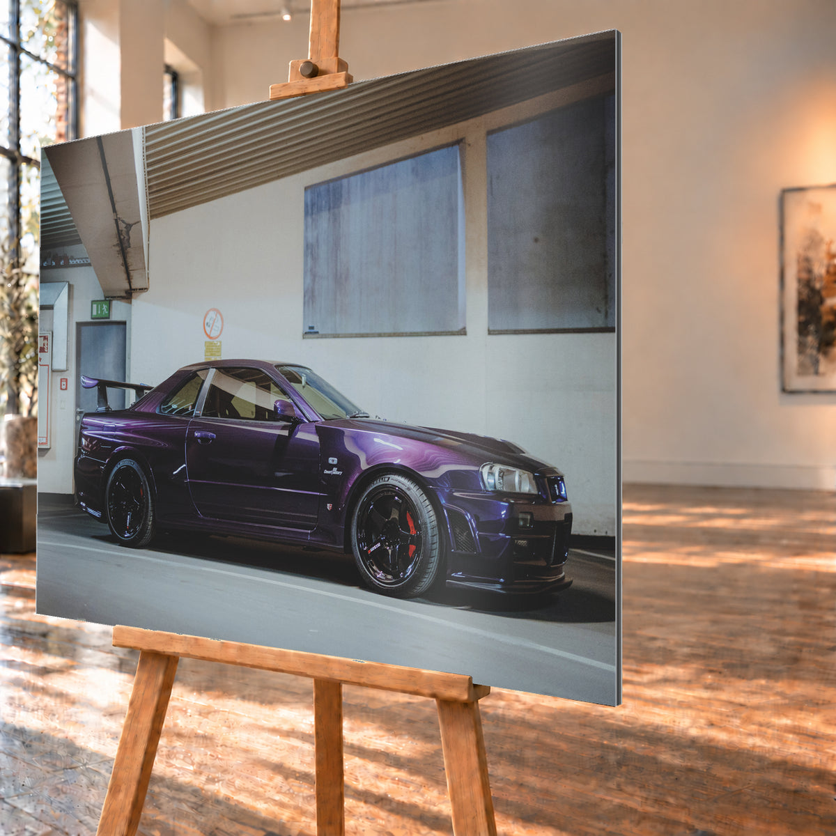 Violetter Nissan Skyline R34, nahezu seitlich mit der Front nach rechts, in einem hellen Parkgaragen-Innenraum; sichtbar sind schwarze Räder, rote Bremssättel und ein großer Heckflügel; Visualisierung als Alu gebürstet-Wandbild, schräg von der Seite auf einer Holzstaffelei.