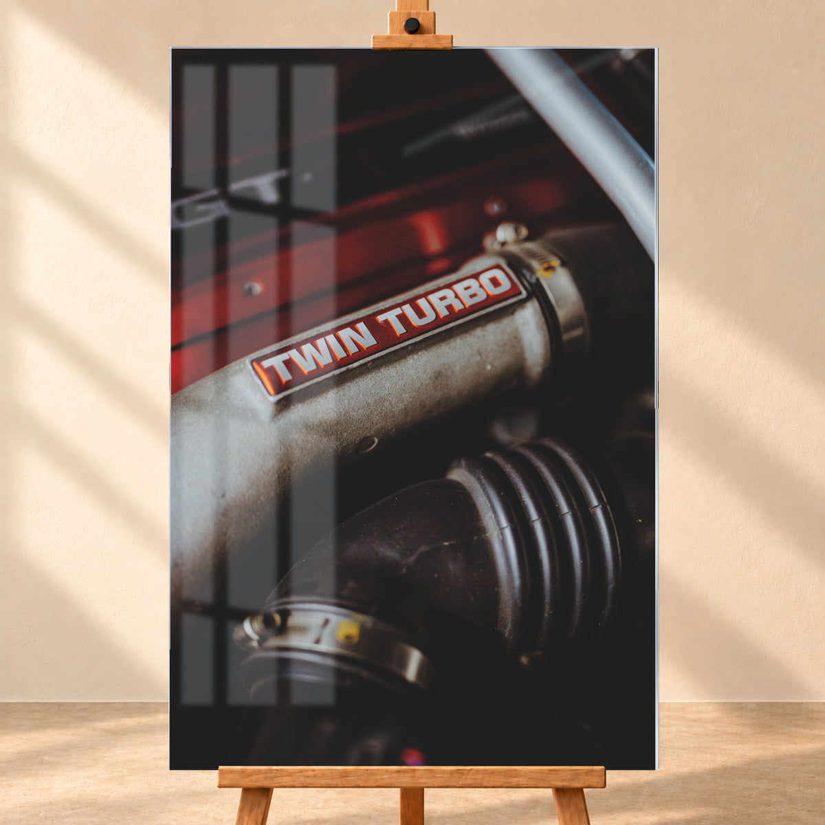 Detailaufnahme eines Motorraums mit metallisch grauem Bauteil und rot-weißem „TWIN TURBO“-Schriftzug. Schwarze gerippte Schläuche, rote Flächen, ein helles diagonales Element und geringe Schärfentiefe prägen das dunkle Bild; Visualisierung als Acrylglas-Wandbild, frontal auf einer Holzstaffelei.