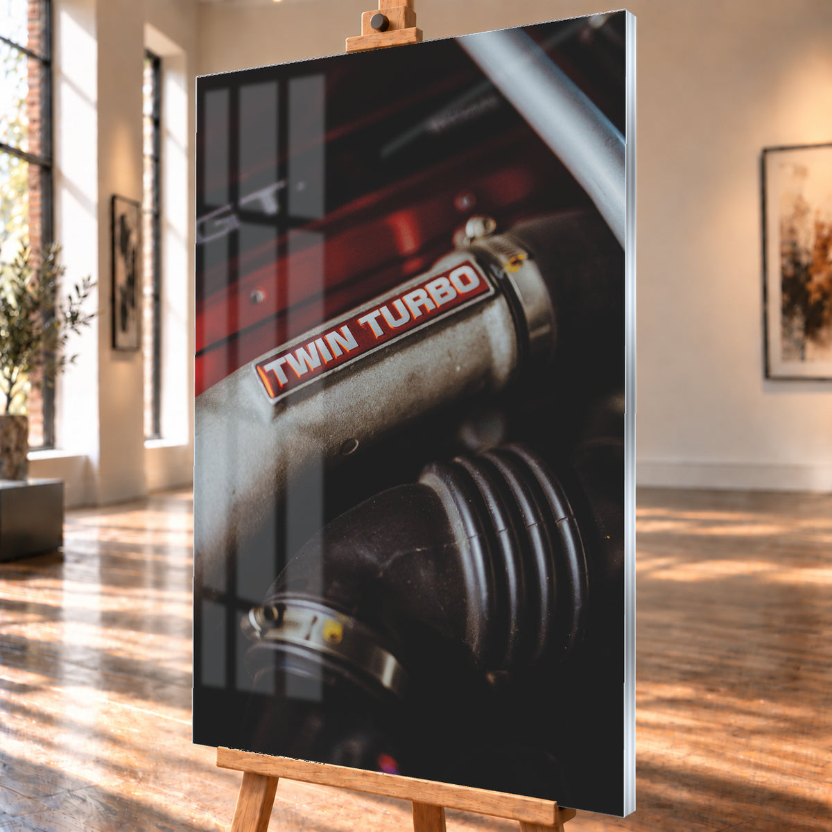 Detailaufnahme eines Motorraums mit metallisch grauem Bauteil und rot-weißem „TWIN TURBO“-Schriftzug. Schwarze gerippte Schläuche, rote Flächen, ein helles diagonales Element und geringe Schärfentiefe prägen das dunkle Bild; Visualisierung als Acrylglas-Wandbild, schräg von der Seite auf einer Holzstaffelei.