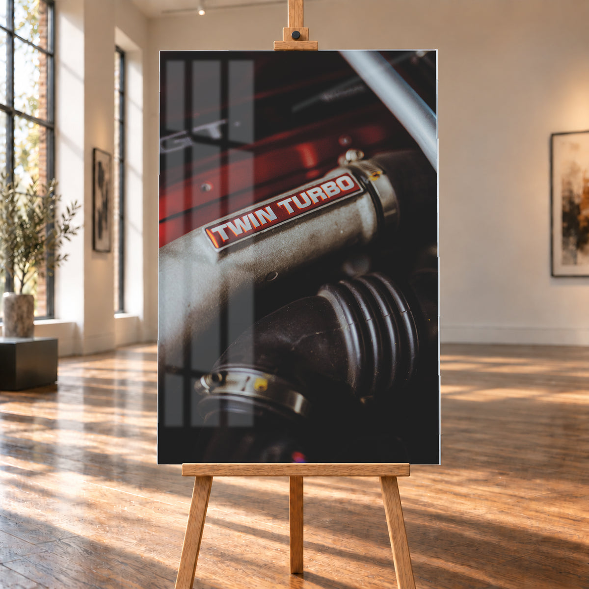 Detailaufnahme eines Motorraums mit metallisch grauem Bauteil und rot-weißem „TWIN TURBO“-Schriftzug. Schwarze gerippte Schläuche, rote Flächen, ein helles diagonales Element und geringe Schärfentiefe prägen das dunkle Bild; Visualisierung als Acrylglas-Wandbild, auf einer Holzstaffelei in einem hellen Galerieraum.