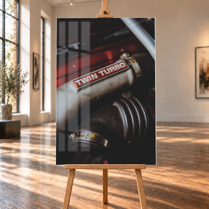 Detailaufnahme eines Motorraums mit metallisch grauem Bauteil und rot-weißem „TWIN TURBO“-Schriftzug. Schwarze gerippte Schläuche, rote Flächen, ein helles diagonales Element und geringe Schärfentiefe prägen das dunkle Bild; Visualisierung als Acrylglas-Wandbild, auf einer Holzstaffelei in einem hellen Galerieraum.