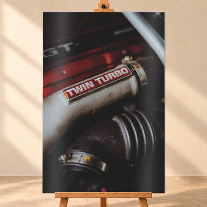 Detailaufnahme eines Motorraums mit metallisch grauem Bauteil und rot-weißem „TWIN TURBO“-Schriftzug. Schwarze gerippte Schläuche, rote Flächen, ein helles diagonales Element und geringe Schärfentiefe prägen das dunkle Bild; Visualisierung als Alu gebürstet-Wandbild, frontal auf einer Holzstaffelei.