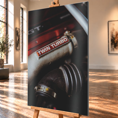 Detailaufnahme eines Motorraums mit metallisch grauem Bauteil und rot-weißem „TWIN TURBO“-Schriftzug. Schwarze gerippte Schläuche, rote Flächen, ein helles diagonales Element und geringe Schärfentiefe prägen das dunkle Bild; Visualisierung als Alu gebürstet-Wandbild, schräg von der Seite auf einer Holzstaffelei.