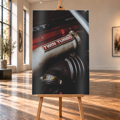 Detailaufnahme eines Motorraums mit metallisch grauem Bauteil und rot-weißem „TWIN TURBO“-Schriftzug. Schwarze gerippte Schläuche, rote Flächen, ein helles diagonales Element und geringe Schärfentiefe prägen das dunkle Bild; Visualisierung als Alu gebürstet-Wandbild, auf einer Holzstaffelei in einem hellen Galerieraum.