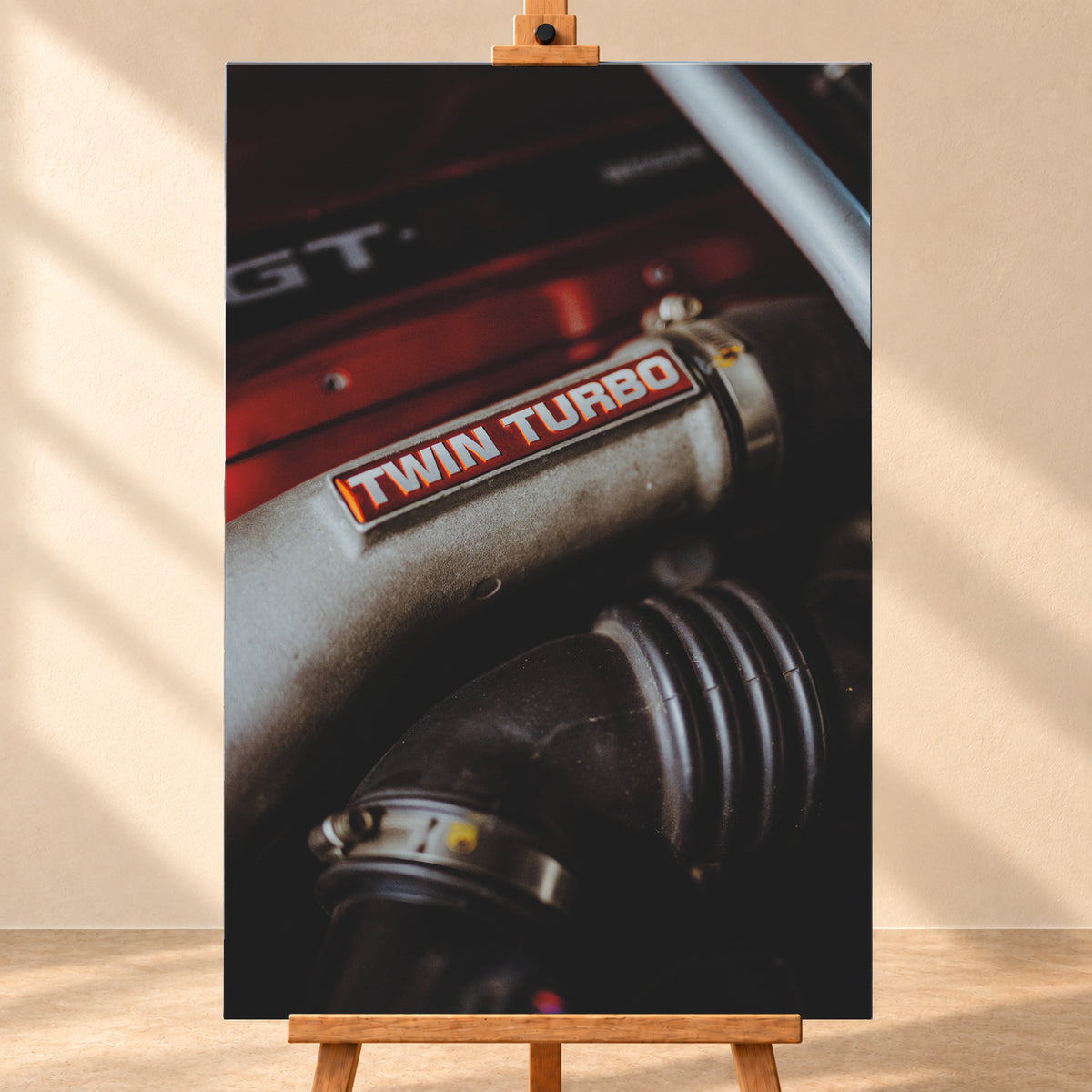 Detailaufnahme eines Motorraums mit metallisch grauem Bauteil und rot-weißem „TWIN TURBO“-Schriftzug. Schwarze gerippte Schläuche, rote Flächen, ein helles diagonales Element und geringe Schärfentiefe prägen das dunkle Bild; Visualisierung als Leinwand-Wandbild, frontal auf einer Holzstaffelei.