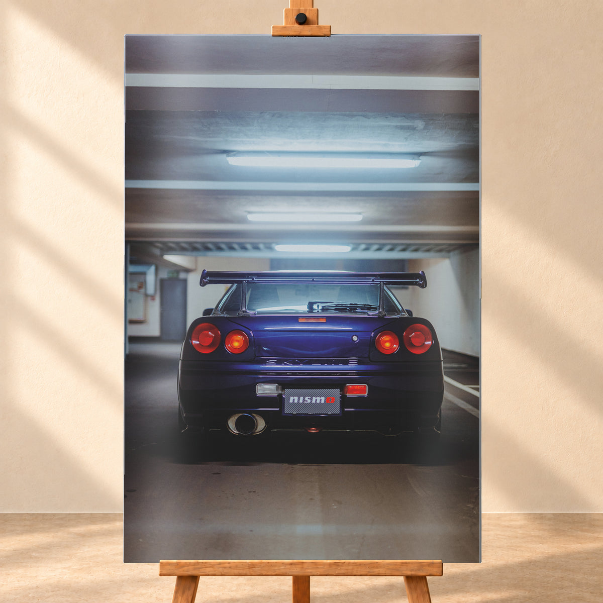 Mittige Heckansicht eines dunkelvioletten Nissan Skyline R34 in einer Parkgarage. Vier runde rote Rückleuchten, davon die beiden inneren orange leuchtend, ein großer Heckflügel, ein rundes Auspuffendrohr und ein Nismo-Schriftzug sind unter kühlen, horizontalen Deckenleuchten zu sehen; Visualisierung als Alu gebürstet-Wandbild, frontal auf einer Holzstaffelei.