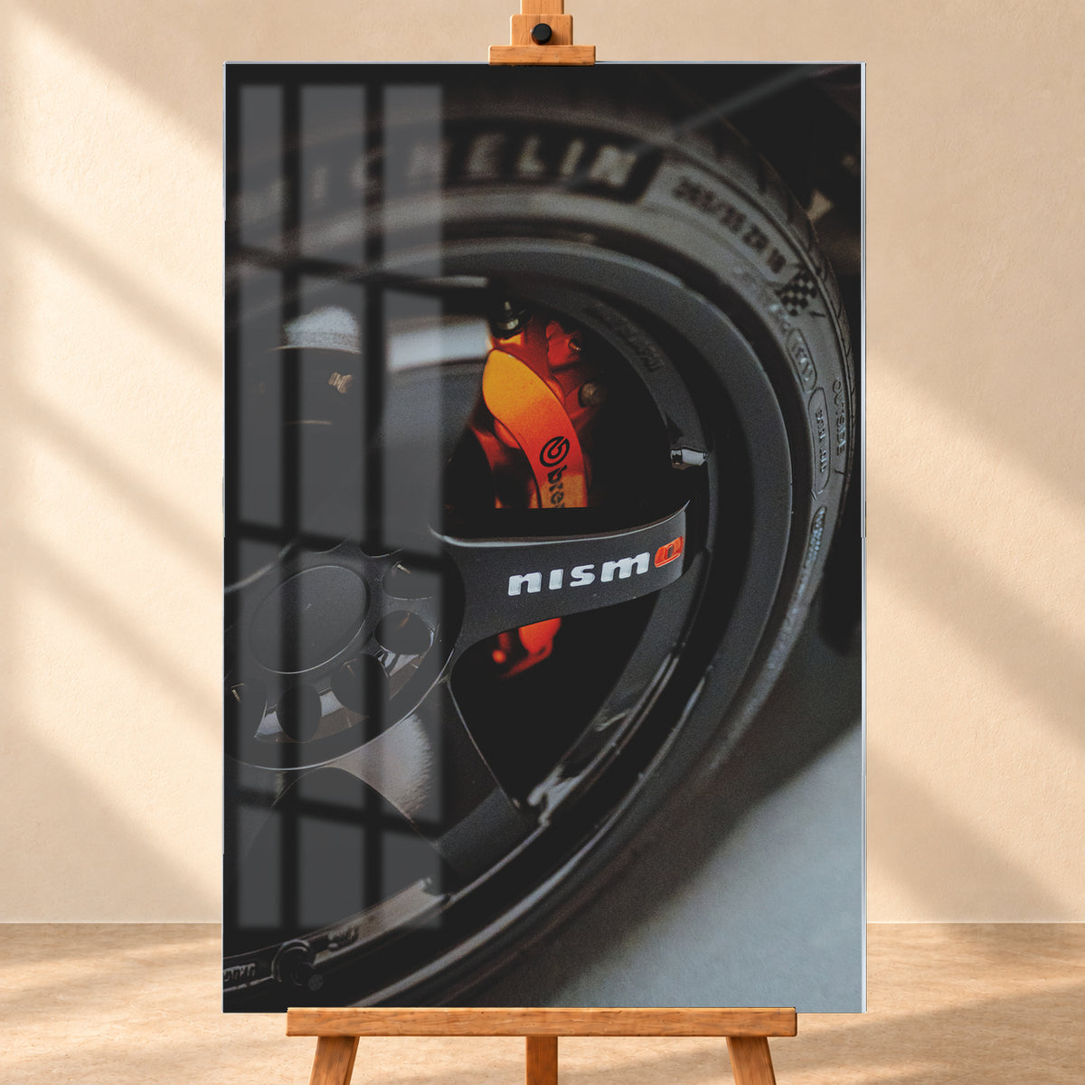 Nahaufnahme einer schwarzen Nismo-Mehrspeichenfelge mit orange-rotem Brembo-Bremssattel und dunkler Reifenflanke; Visualisierung als Acrylglas-Wandbild, frontal auf einer Holzstaffelei.