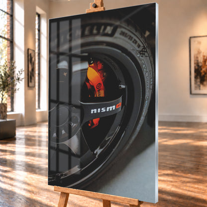 Nahaufnahme einer schwarzen Nismo-Mehrspeichenfelge mit orange-rotem Brembo-Bremssattel und dunkler Reifenflanke; Visualisierung als Acrylglas-Wandbild, schräg von der Seite auf einer Holzstaffelei.