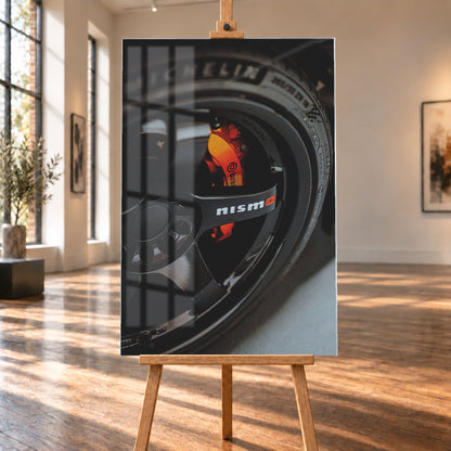 Nahaufnahme einer schwarzen Nismo-Mehrspeichenfelge mit orange-rotem Brembo-Bremssattel und dunkler Reifenflanke; Visualisierung als Acrylglas-Wandbild, auf einer Holzstaffelei in einem hellen Galerieraum.