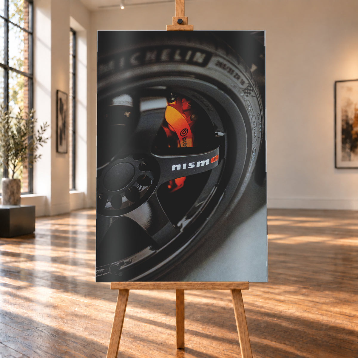 Nahaufnahme einer schwarzen Nismo-Mehrspeichenfelge mit orange-rotem Brembo-Bremssattel und dunkler Reifenflanke; Visualisierung als Alu gebürstet-Wandbild, auf einer Holzstaffelei in einem hellen Galerieraum.