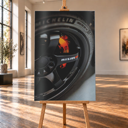 Nahaufnahme einer schwarzen Nismo-Mehrspeichenfelge mit orange-rotem Brembo-Bremssattel und dunkler Reifenflanke; Visualisierung als Alu gebürstet-Wandbild, auf einer Holzstaffelei in einem hellen Galerieraum.