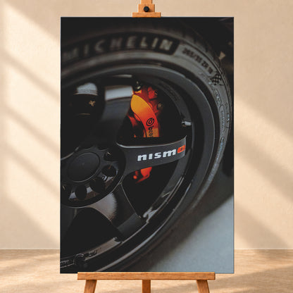 Nahaufnahme einer schwarzen Nismo-Mehrspeichenfelge mit orange-rotem Brembo-Bremssattel und dunkler Reifenflanke; Visualisierung als Leinwand-Wandbild, frontal auf einer Holzstaffelei.