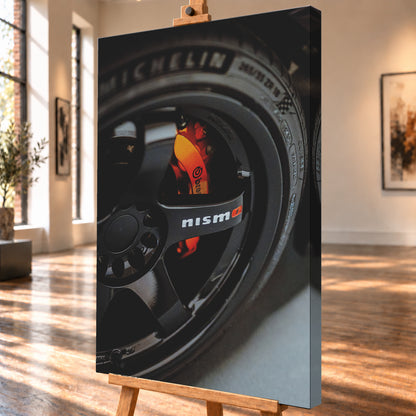 Nahaufnahme einer schwarzen Nismo-Mehrspeichenfelge mit orange-rotem Brembo-Bremssattel und dunkler Reifenflanke; Visualisierung als Leinwand-Wandbild, schräg von der Seite auf einer Holzstaffelei.