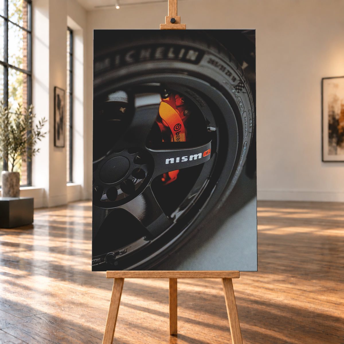 Nahaufnahme einer schwarzen Nismo-Mehrspeichenfelge mit orange-rotem Brembo-Bremssattel und dunkler Reifenflanke; Visualisierung als Leinwand-Wandbild, auf einer Holzstaffelei in einem hellen Galerieraum.