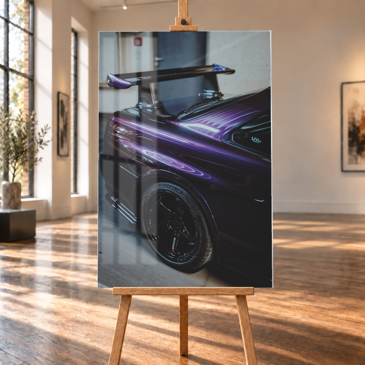 Nahaufnahme der hinteren Seitenpartie eines violetten Nissan Skyline R34 in einem Innenraum; sichtbar sind schwarzes Hinterrad, Radlauf, Heckscheibe und großer Heckflügel mit hellen Reflexionen; Visualisierung als Acrylglas-Wandbild, auf einer Holzstaffelei in einem hellen Galerieraum.
