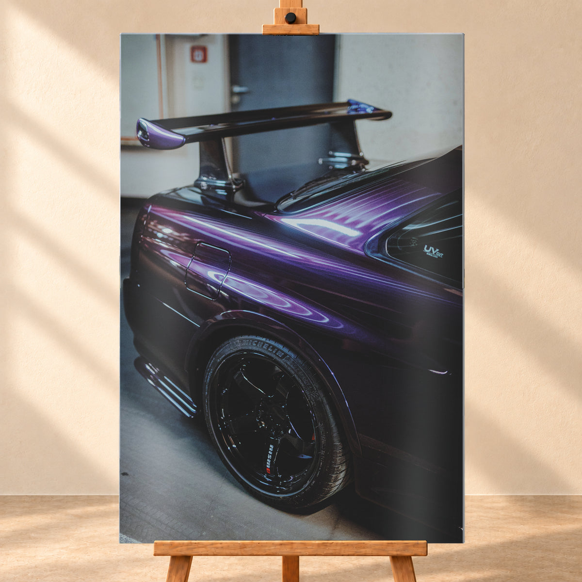Nahaufnahme der hinteren Seitenpartie eines violetten Nissan Skyline R34 in einem Innenraum; sichtbar sind schwarzes Hinterrad, Radlauf, Heckscheibe und großer Heckflügel mit hellen Reflexionen; Visualisierung als Alu gebürstet-Wandbild, frontal auf einer Holzstaffelei.
