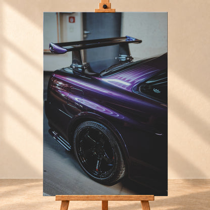 Nahaufnahme der hinteren Seitenpartie eines violetten Nissan Skyline R34 in einem Innenraum; sichtbar sind schwarzes Hinterrad, Radlauf, Heckscheibe und großer Heckflügel mit hellen Reflexionen; Visualisierung als Leinwand-Wandbild, frontal auf einer Holzstaffelei.