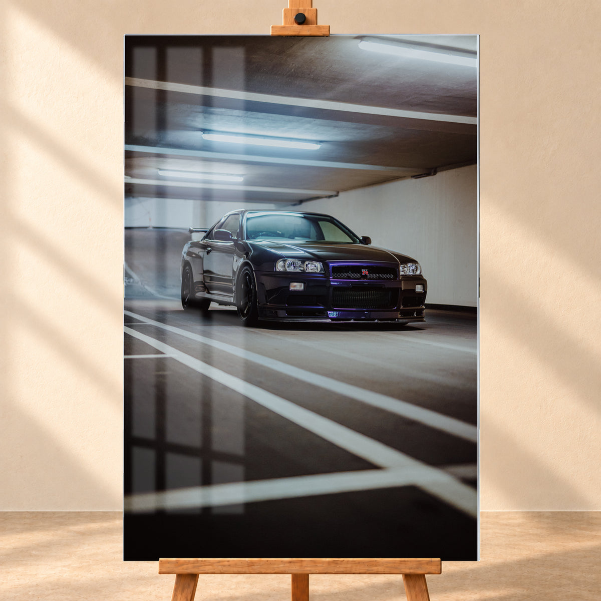 Dunkler Nissan Skyline R34 GT-R in niedriger Front-Dreiviertelansicht in einer Beton-Parkgarage, mit violett-blauen Reflexen, dunklen Rädern, Heckflügel, weißen Fahrbahnlinien und kühlen Deckenleuchten; Visualisierung als Acrylglas-Wandbild, frontal auf einer Holzstaffelei.