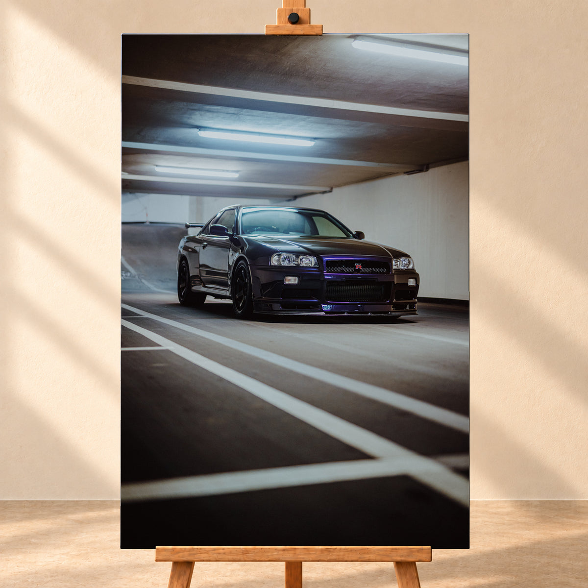 Dunkler Nissan Skyline R34 GT-R in niedriger Front-Dreiviertelansicht in einer Beton-Parkgarage, mit violett-blauen Reflexen, dunklen Rädern, Heckflügel, weißen Fahrbahnlinien und kühlen Deckenleuchten; Visualisierung als Leinwand-Wandbild, frontal auf einer Holzstaffelei.