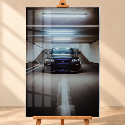 Dunkler Nissan Skyline R34 in frontaler Ansicht auf einer Betonrampe, unter kühlen weißen Deckenleuchten, mit mittiger Fahrbahnmarkierung und violettblauen Reflexen an Kühlergrill und Frontschürze; Visualisierung als Acrylglas-Wandbild, frontal auf einer Holzstaffelei.