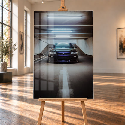 Dunkler Nissan Skyline R34 in frontaler Ansicht auf einer Betonrampe, unter kühlen weißen Deckenleuchten, mit mittiger Fahrbahnmarkierung und violettblauen Reflexen an Kühlergrill und Frontschürze; Visualisierung als Acrylglas-Wandbild, auf einer Holzstaffelei in einem hellen Galerieraum.