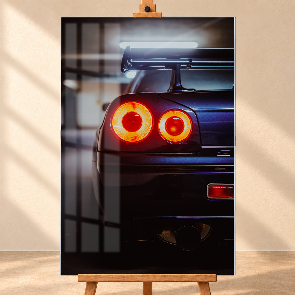 Nahaufnahme der Heckpartie eines dunklen Nissan Skyline R34 V-Spec mit zwei sichtbaren, rot-orange leuchtenden Rundrückleuchten, großem Heckflügel und rundem Auspuffendrohr vor unscharfem, kühl beleuchtetem Hintergrund; Visualisierung als Acrylglas-Wandbild, frontal auf einer Holzstaffelei.