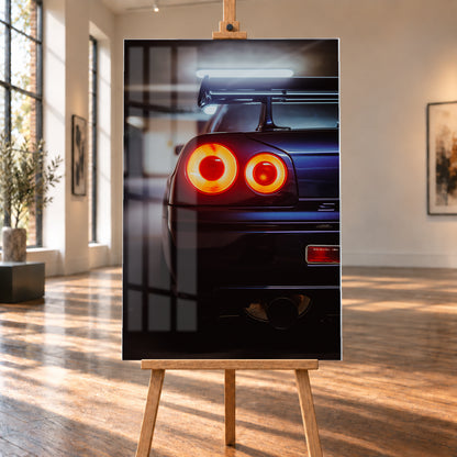 Nahaufnahme der Heckpartie eines dunklen Nissan Skyline R34 V-Spec mit zwei sichtbaren, rot-orange leuchtenden Rundrückleuchten, großem Heckflügel und rundem Auspuffendrohr vor unscharfem, kühl beleuchtetem Hintergrund; Visualisierung als Acrylglas-Wandbild, auf einer Holzstaffelei in einem hellen Galerieraum.