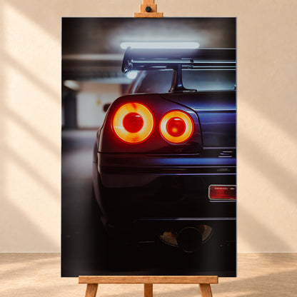 Nahaufnahme der Heckpartie eines dunklen Nissan Skyline R34 V-Spec mit zwei sichtbaren, rot-orange leuchtenden Rundrückleuchten, großem Heckflügel und rundem Auspuffendrohr vor unscharfem, kühl beleuchtetem Hintergrund; Visualisierung als Alu gebürstet-Wandbild, frontal auf einer Holzstaffelei.