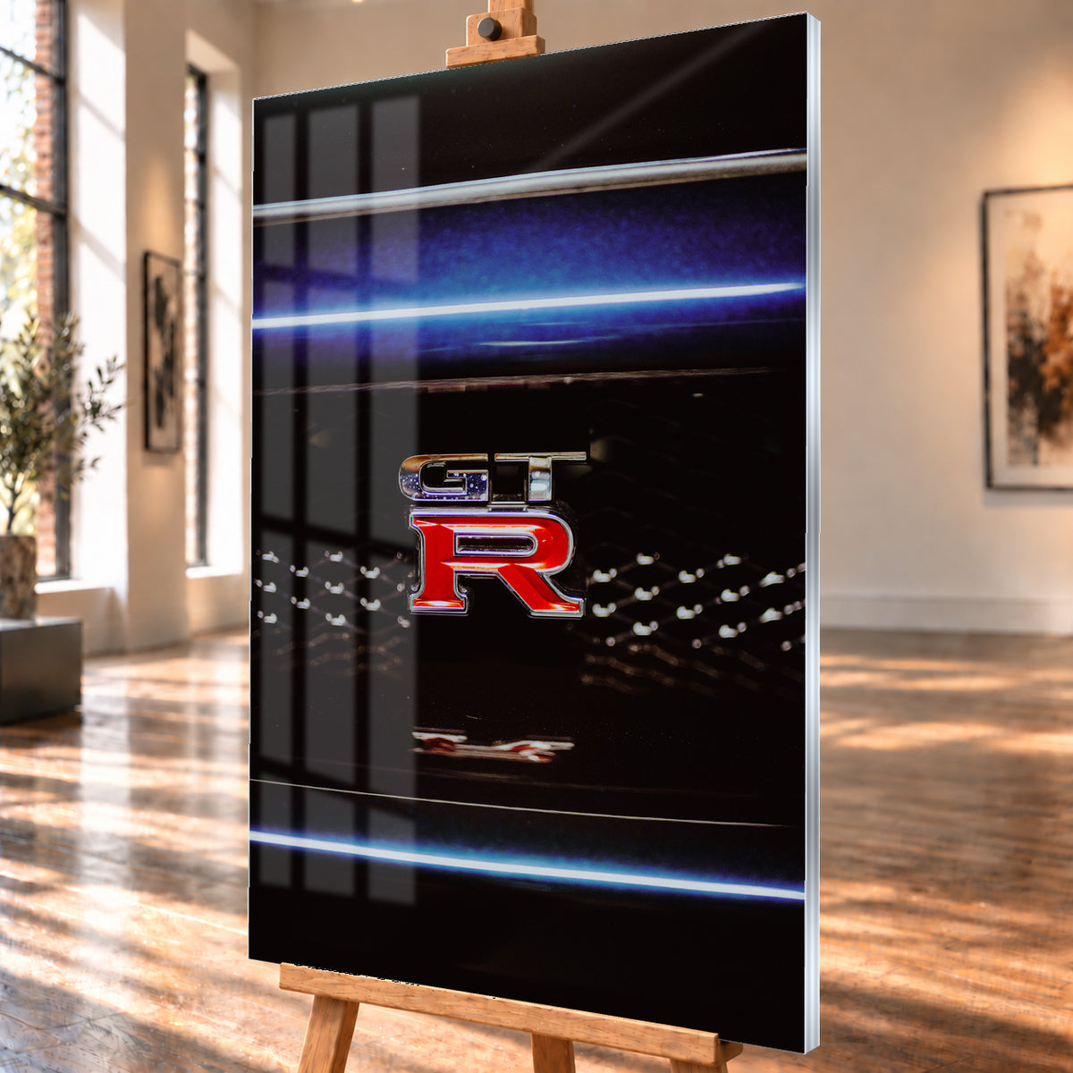 Hochformatige Nahaufnahme eines rot-silbernen GT-R-Emblems auf einem schwarzen Wabengrill. Blaue horizontale Lichtbänder verlaufen ober- und unterhalb; kleine helle Reflexe glitzern in der dunklen Fläche; Visualisierung als Acrylglas-Wandbild, schräg von der Seite auf einer Holzstaffelei.