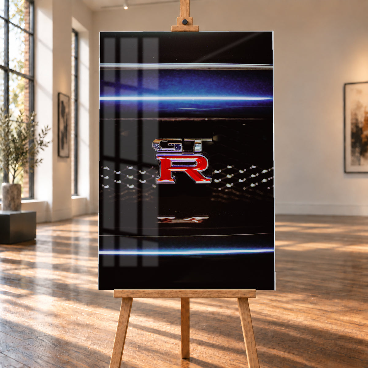 Hochformatige Nahaufnahme eines rot-silbernen GT-R-Emblems auf einem schwarzen Wabengrill. Blaue horizontale Lichtbänder verlaufen ober- und unterhalb; kleine helle Reflexe glitzern in der dunklen Fläche; Visualisierung als Acrylglas-Wandbild, auf einer Holzstaffelei in einem hellen Galerieraum.