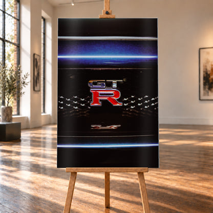 Hochformatige Nahaufnahme eines rot-silbernen GT-R-Emblems auf einem schwarzen Wabengrill. Blaue horizontale Lichtbänder verlaufen ober- und unterhalb; kleine helle Reflexe glitzern in der dunklen Fläche; Visualisierung als Alu gebürstet-Wandbild, auf einer Holzstaffelei in einem hellen Galerieraum.