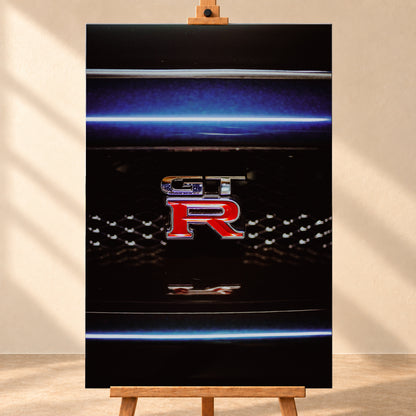 Hochformatige Nahaufnahme eines rot-silbernen GT-R-Emblems auf einem schwarzen Wabengrill. Blaue horizontale Lichtbänder verlaufen ober- und unterhalb; kleine helle Reflexe glitzern in der dunklen Fläche; Visualisierung als Leinwand-Wandbild, frontal auf einer Holzstaffelei.