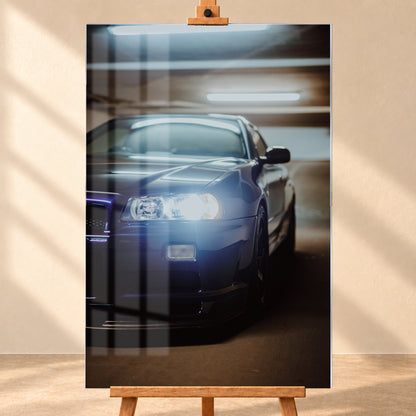Hochformatige Nahaufnahme eines dunkelblau bis schwarz wirkenden Nissan Skyline R34 in schräger Frontansicht. Ein heller Frontscheinwerfer leuchtet in die Kamera, während sich kühle rechteckige Deckenlichter auf Motorhaube und Windschutzscheibe spiegeln. Das Fahrzeug steht in einem warm beleuchteten, unscharfen Innenraum; Visualisierung als Acrylglas-Wandbild, frontal auf einer Holzstaffelei.