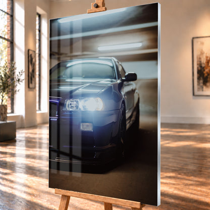 Hochformatige Nahaufnahme eines dunkelblau bis schwarz wirkenden Nissan Skyline R34 in schräger Frontansicht. Ein heller Frontscheinwerfer leuchtet in die Kamera, während sich kühle rechteckige Deckenlichter auf Motorhaube und Windschutzscheibe spiegeln. Das Fahrzeug steht in einem warm beleuchteten, unscharfen Innenraum; Visualisierung als Acrylglas-Wandbild, schräg von der Seite auf einer Holzstaffelei.