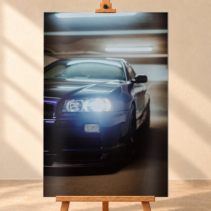 Hochformatige Nahaufnahme eines dunkelblau bis schwarz wirkenden Nissan Skyline R34 in schräger Frontansicht. Ein heller Frontscheinwerfer leuchtet in die Kamera, während sich kühle rechteckige Deckenlichter auf Motorhaube und Windschutzscheibe spiegeln. Das Fahrzeug steht in einem warm beleuchteten, unscharfen Innenraum; Visualisierung als Alu gebürstet-Wandbild, frontal auf einer Holzstaffelei.