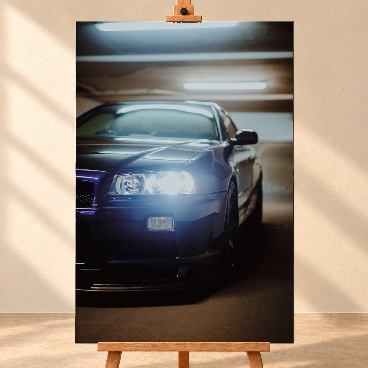 Hochformatige Nahaufnahme eines dunkelblau bis schwarz wirkenden Nissan Skyline R34 in schräger Frontansicht. Ein heller Frontscheinwerfer leuchtet in die Kamera, während sich kühle rechteckige Deckenlichter auf Motorhaube und Windschutzscheibe spiegeln. Das Fahrzeug steht in einem warm beleuchteten, unscharfen Innenraum; Visualisierung als Leinwand-Wandbild, frontal auf einer Holzstaffelei.