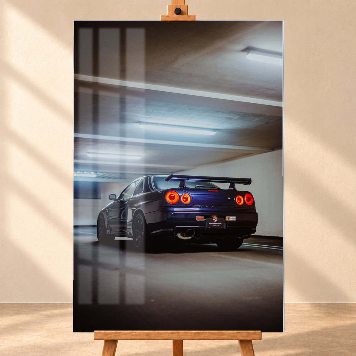 Dunkler Nissan Skyline R34 in niedriger Heck-Dreiviertelansicht mit vier rot leuchtenden Rundrückleuchten, großem Heckflügel und sichtbarem Auspuff in einer Beton-Parkgarage unter kühlen Deckenleuchten; Visualisierung als Acrylglas-Wandbild, frontal auf einer Holzstaffelei.