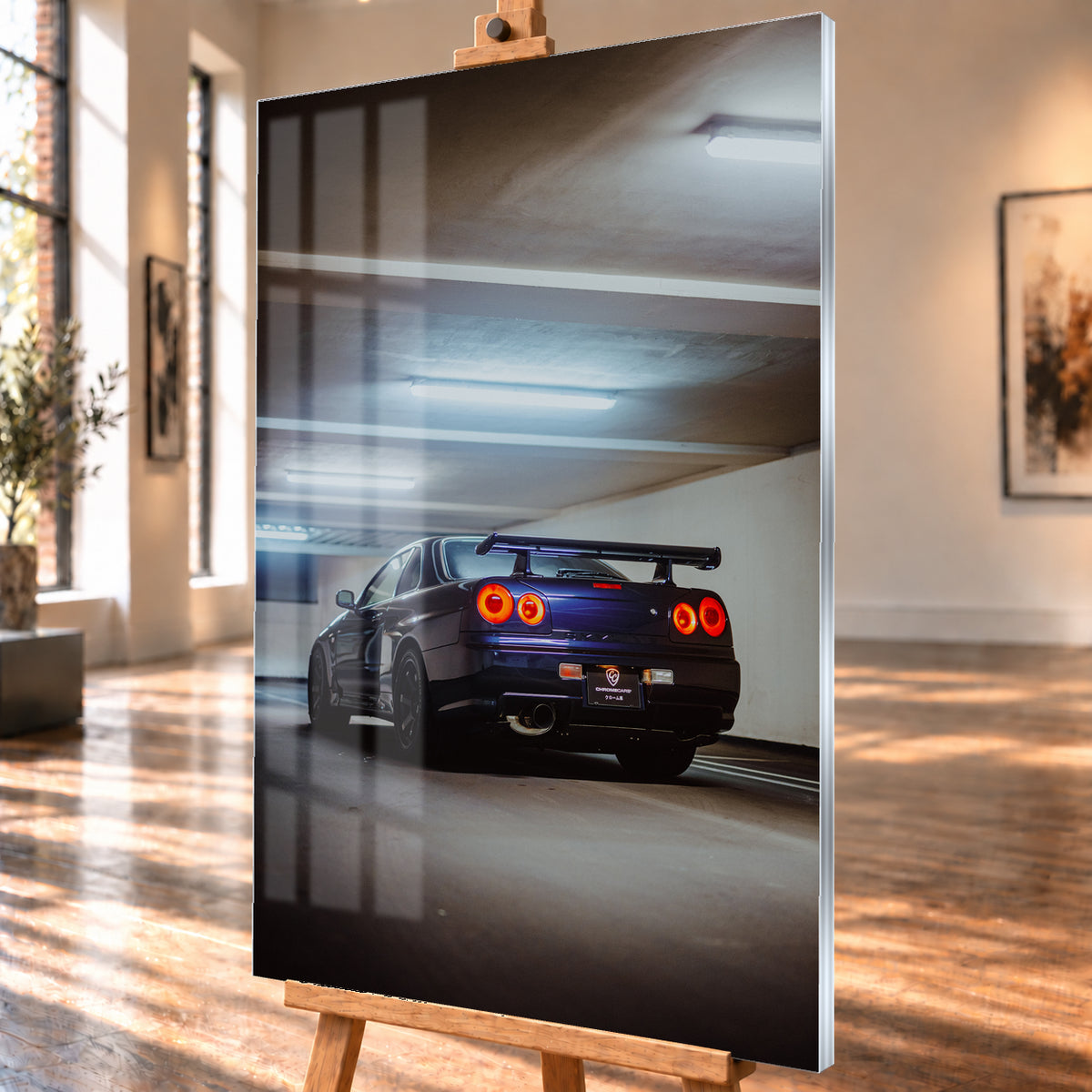 Dunkler Nissan Skyline R34 in niedriger Heck-Dreiviertelansicht mit vier rot leuchtenden Rundrückleuchten, großem Heckflügel und sichtbarem Auspuff in einer Beton-Parkgarage unter kühlen Deckenleuchten; Visualisierung als Acrylglas-Wandbild, schräg von der Seite auf einer Holzstaffelei.