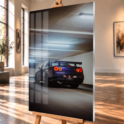Dunkler Nissan Skyline R34 in niedriger Heck-Dreiviertelansicht mit vier rot leuchtenden Rundrückleuchten, großem Heckflügel und sichtbarem Auspuff in einer Beton-Parkgarage unter kühlen Deckenleuchten; Visualisierung als Acrylglas-Wandbild, schräg von der Seite auf einer Holzstaffelei.