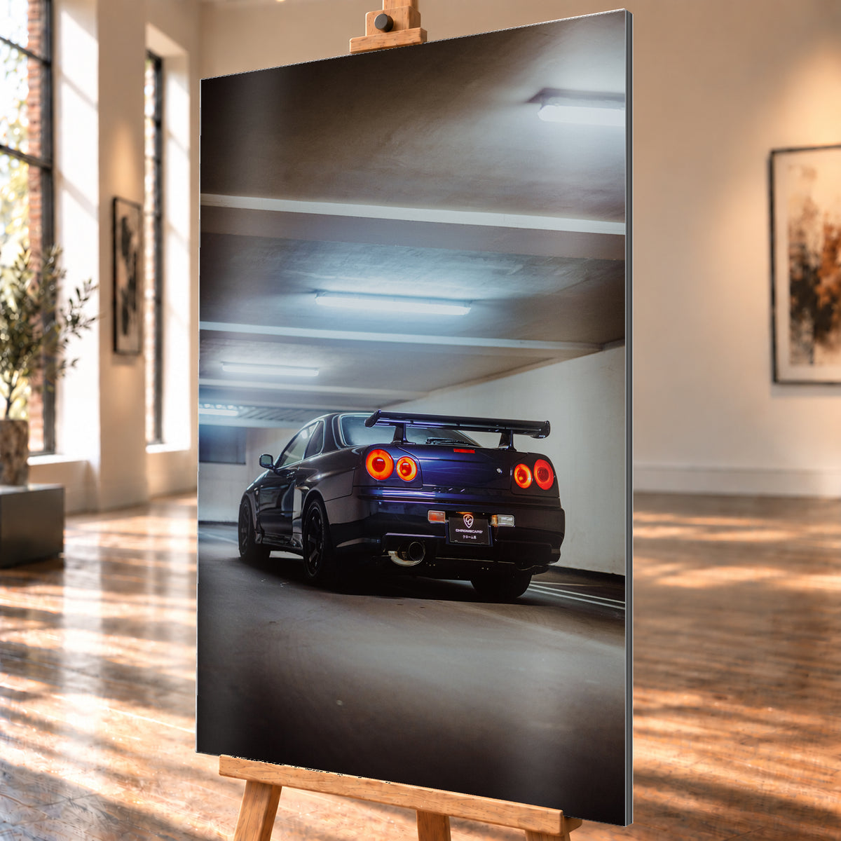 Dunkler Nissan Skyline R34 in niedriger Heck-Dreiviertelansicht mit vier rot leuchtenden Rundrückleuchten, großem Heckflügel und sichtbarem Auspuff in einer Beton-Parkgarage unter kühlen Deckenleuchten; Visualisierung als Alu gebürstet-Wandbild, schräg von der Seite auf einer Holzstaffelei.