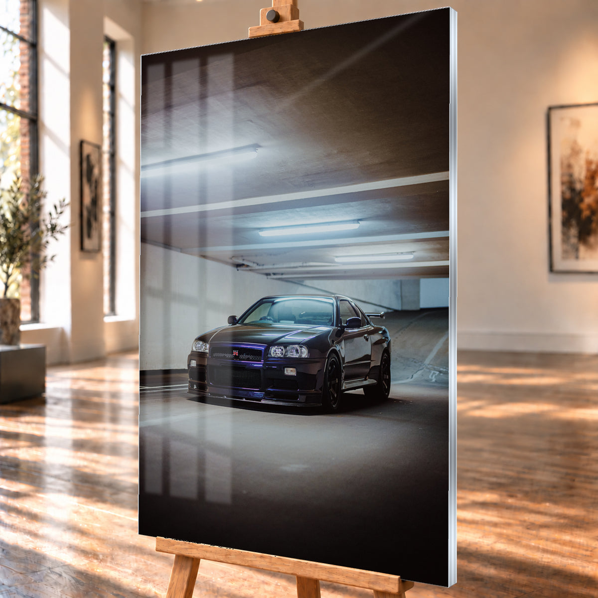 Dunkel lackierter Nissan Skyline R34 in frontaler Dreiviertelansicht in einer Betongarage mit weißen Deckenleuchten, Fahrbahnmarkierungen und ansteigender Rampe im Hintergrund; Visualisierung als Acrylglas-Wandbild, schräg von der Seite auf einer Holzstaffelei.