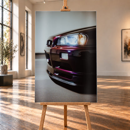 Nahaufnahme der Front eines dunklen, bordeauxviolett wirkenden Nissan Skyline V-Spec aus niedriger, schräger Perspektive. Ein rechteckiger Scheinwerfer mit kühlen und warmen Lichtanteilen dominiert die rechte Bildhälfte. Darunter sind Frontschürze, Leuchte und Lufteinlass zu sehen; eine hellgraue Wand und ein grauer Boden bilden den unscharfen Hintergrund; Visualisierung als Alu gebürstet-Wandbild, auf einer Holzstaffelei in einem hellen Galerieraum.