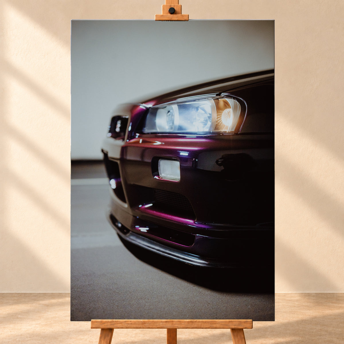 Nahaufnahme der Front eines dunklen, bordeauxviolett wirkenden Nissan Skyline V-Spec aus niedriger, schräger Perspektive. Ein rechteckiger Scheinwerfer mit kühlen und warmen Lichtanteilen dominiert die rechte Bildhälfte. Darunter sind Frontschürze, Leuchte und Lufteinlass zu sehen; eine hellgraue Wand und ein grauer Boden bilden den unscharfen Hintergrund; Visualisierung als Leinwand-Wandbild, frontal auf einer Holzstaffelei.