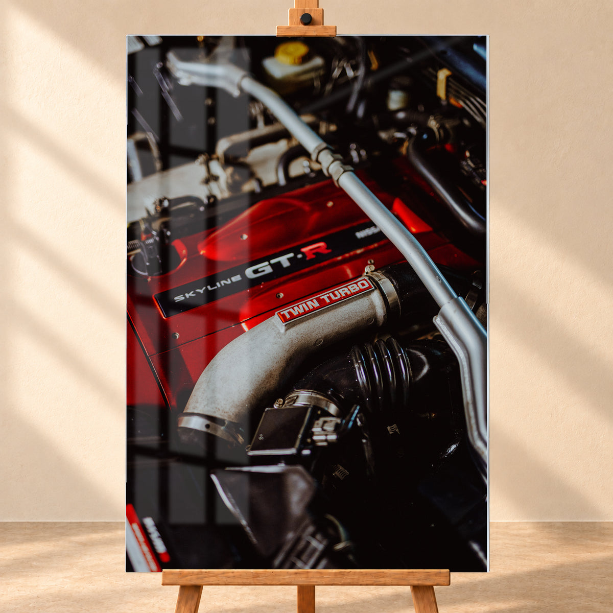 Nahaufnahme eines Motorraums mit roter Abdeckung und sichtbarem SKYLINE GT-R-Schriftzug, hellem Rohr mit TWIN TURBO-Kennzeichnung, schwarzer Verrohrung und einer silbernen, diagonal verlaufenden Strebe; dunkler Hintergrund mit warmen Lichtreflexen; Visualisierung als Acrylglas-Wandbild, frontal auf einer Holzstaffelei.