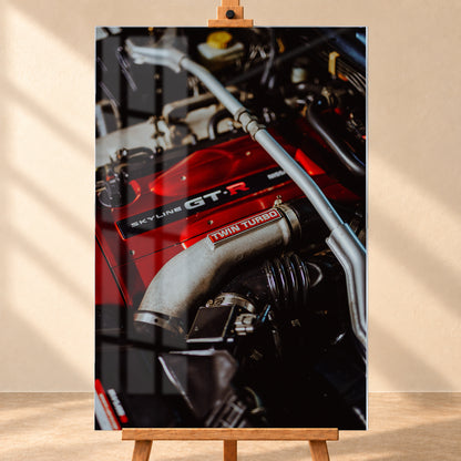 Nahaufnahme eines Motorraums mit roter Abdeckung und sichtbarem SKYLINE GT-R-Schriftzug, hellem Rohr mit TWIN TURBO-Kennzeichnung, schwarzer Verrohrung und einer silbernen, diagonal verlaufenden Strebe; dunkler Hintergrund mit warmen Lichtreflexen; Visualisierung als Acrylglas-Wandbild, frontal auf einer Holzstaffelei.