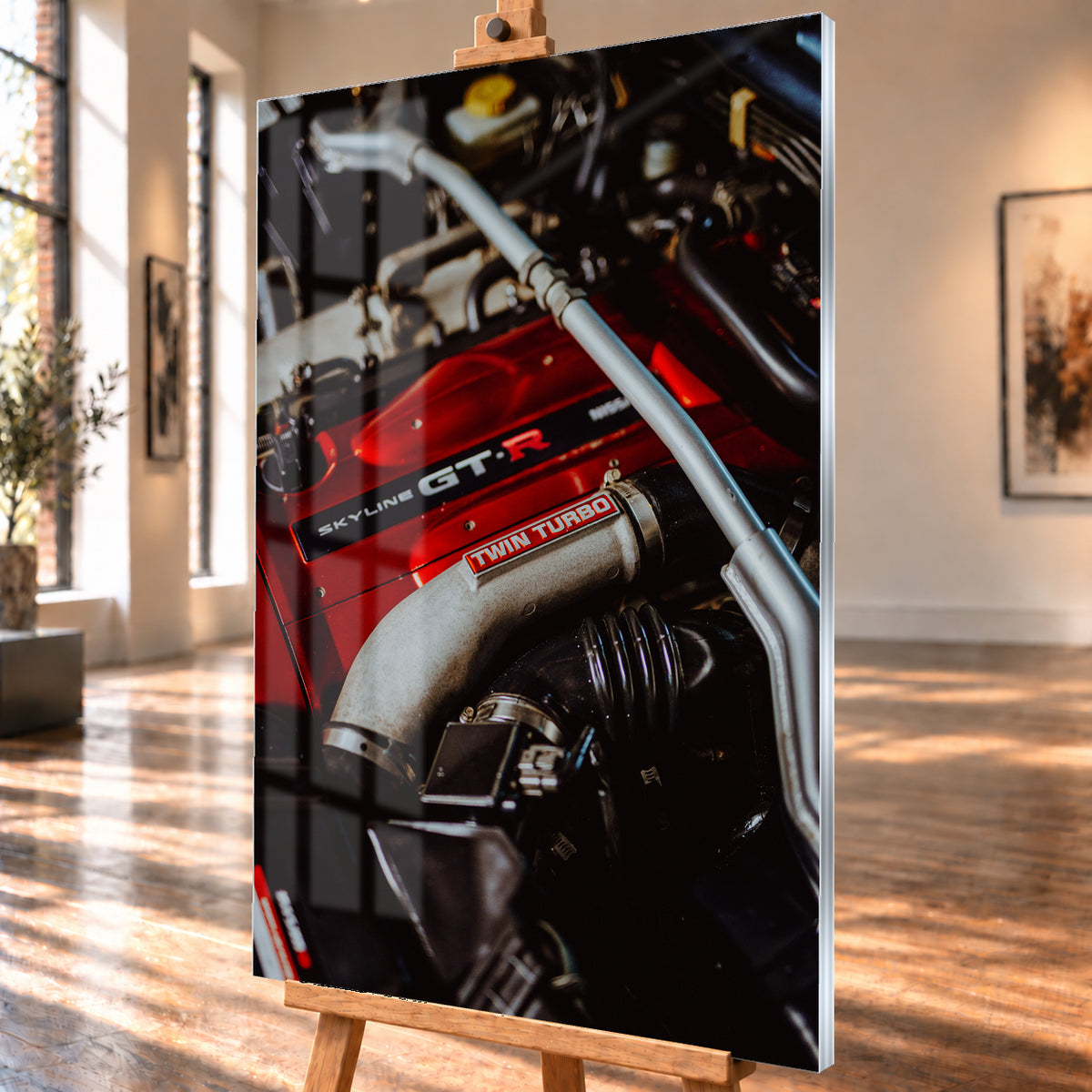 Nahaufnahme eines Motorraums mit roter Abdeckung und sichtbarem SKYLINE GT-R-Schriftzug, hellem Rohr mit TWIN TURBO-Kennzeichnung, schwarzer Verrohrung und einer silbernen, diagonal verlaufenden Strebe; dunkler Hintergrund mit warmen Lichtreflexen; Visualisierung als Acrylglas-Wandbild, schräg von der Seite auf einer Holzstaffelei.