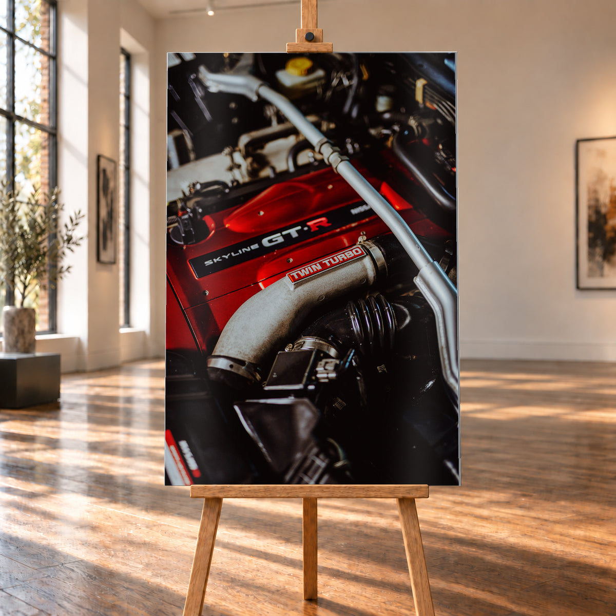Nahaufnahme eines Motorraums mit roter Abdeckung und sichtbarem SKYLINE GT-R-Schriftzug, hellem Rohr mit TWIN TURBO-Kennzeichnung, schwarzer Verrohrung und einer silbernen, diagonal verlaufenden Strebe; dunkler Hintergrund mit warmen Lichtreflexen; Visualisierung als Alu gebürstet-Wandbild, auf einer Holzstaffelei in einem hellen Galerieraum.